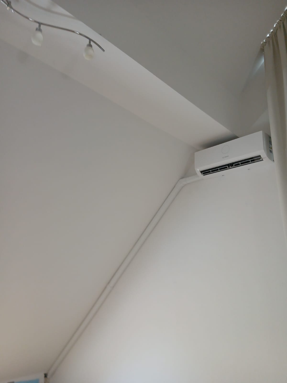 Innenklimagerät an schräger Decke mit sichtbarer Verrohrung und Deckenleuchte im weißen Zimmer.
